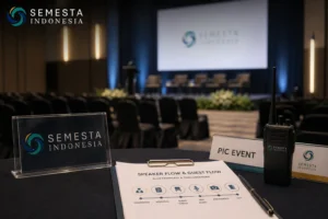 Tampilan meja koordinasi acara formal dengan papan nama Semesta Indonesia, dokumen speaker flow dan guest flow, perangkat komunikasi, serta suasana ruang seminar di latar belakang.